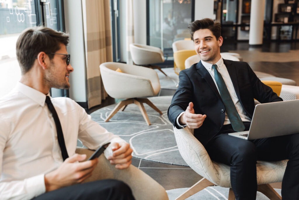 Photo of a pleased businessmen colleagues indoors in business center office using mobile phone and laptop computer.