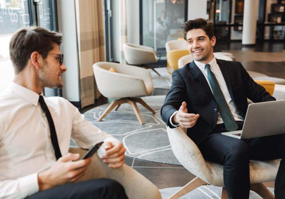 Photo of a pleased businessmen colleagues indoors in business center office using mobile phone and laptop computer.